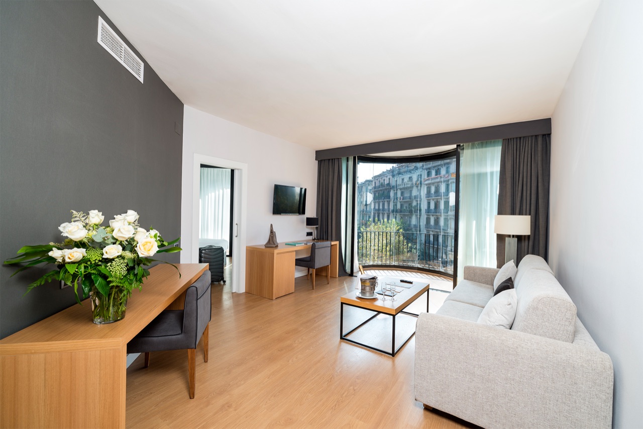 Suite living area with a grey sofa, wooden desk, flowers on a console table and a curved bay window overlooking the street.