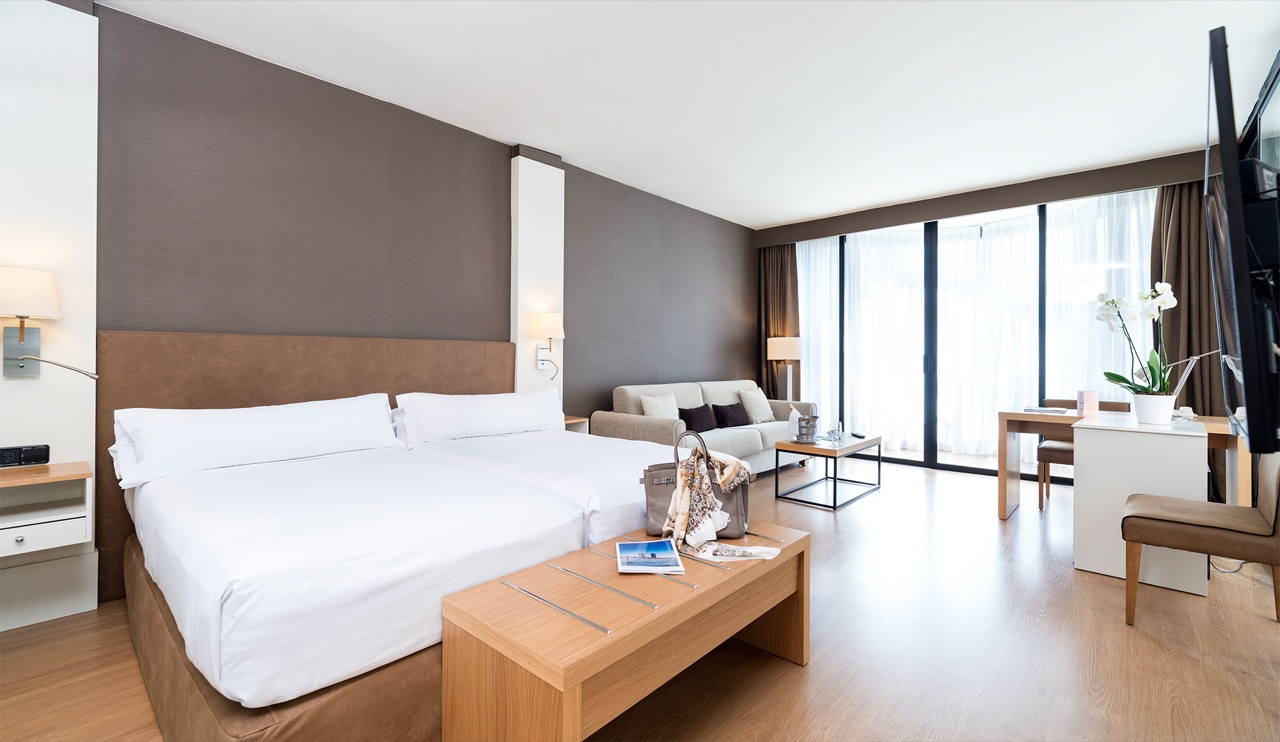 Executive room with two twin beds pushed together with white linens, a wooden bench, a sofa and a desk near the window.