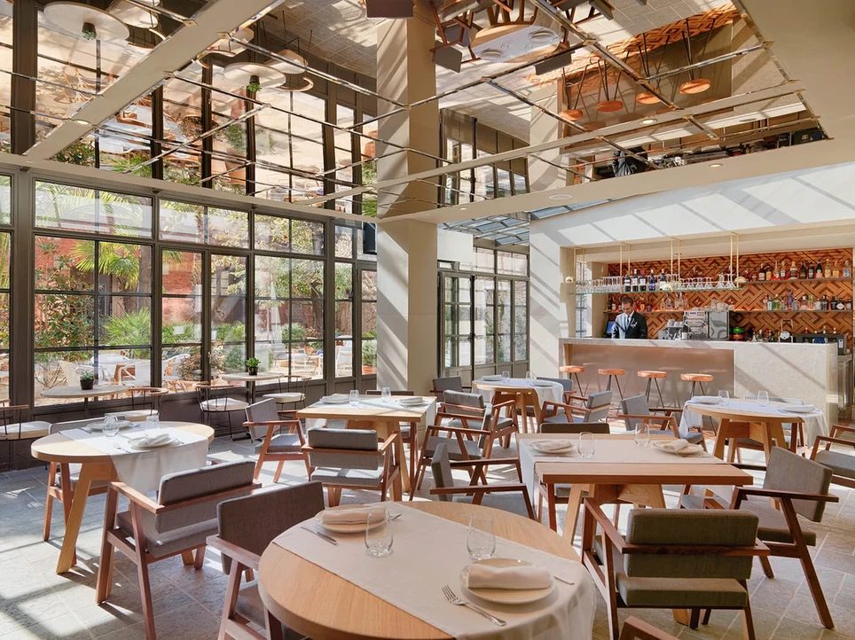 El Til.ler restaurant at H10 Casa Mimosa, a skylit atrium dining room with a mirrored ceiling, wood-and-canvas chairs, and floor-to-ceiling windows onto the garden
