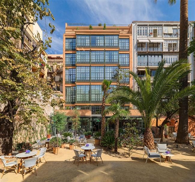 Rear facade of H10 Casa Mimosa seen from the El Til.ler garden patio, with wicker cafe chairs, palm trees, and the building's industrial-era brick and glass windows