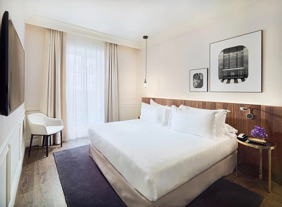 Deluxe room at H10 Casa Mimosa with a white bed against a walnut headboard wall, framed black-and-white photography, and a curtained balcony window