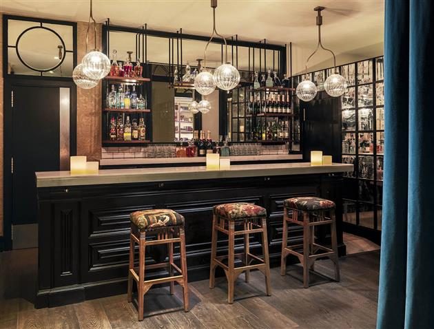 The Library lobby bar at H10 Catalunya Plaza with a dark wood counter, globe pendant lights, a backlit bottle display, and patterned velvet bar stools