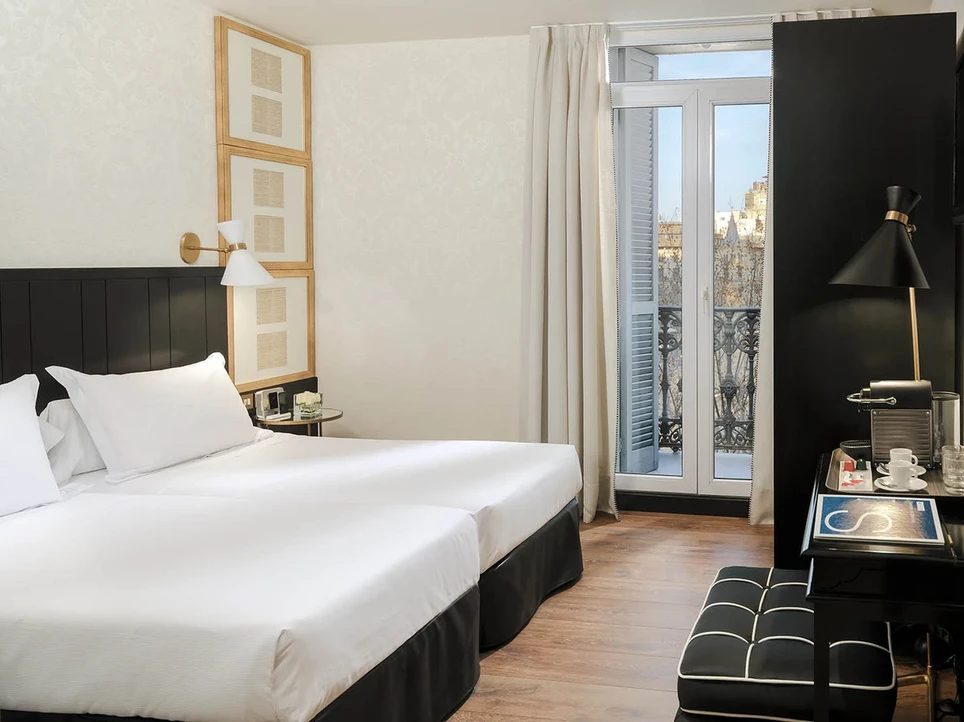 Deluxe room at H10 Catalunya Plaza with twin beds against a black headboard wall and a Juliet balcony overlooking Barcelona's rooftops