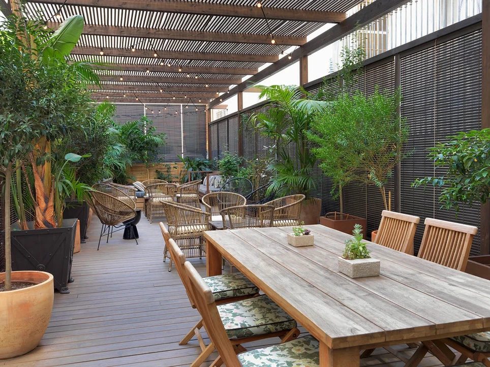 Covered garden terrace at H10 Catalunya Plaza with a wood pergola strung with lights, potted palms, rattan chairs, and a long communal table