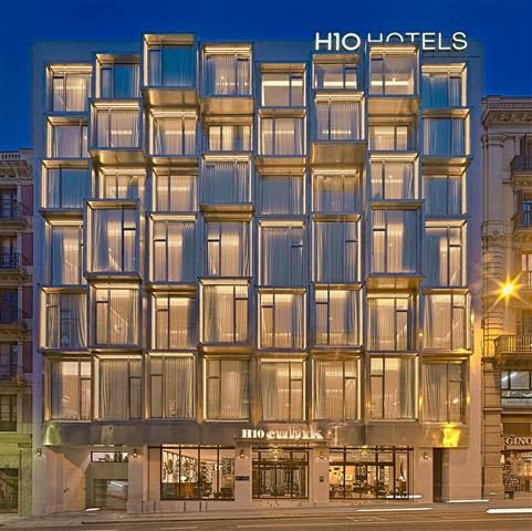 Night view of H10 Cubik's facade of stacked glass-box balconies with the H10 Hotels sign lit above, on a Barcelona street