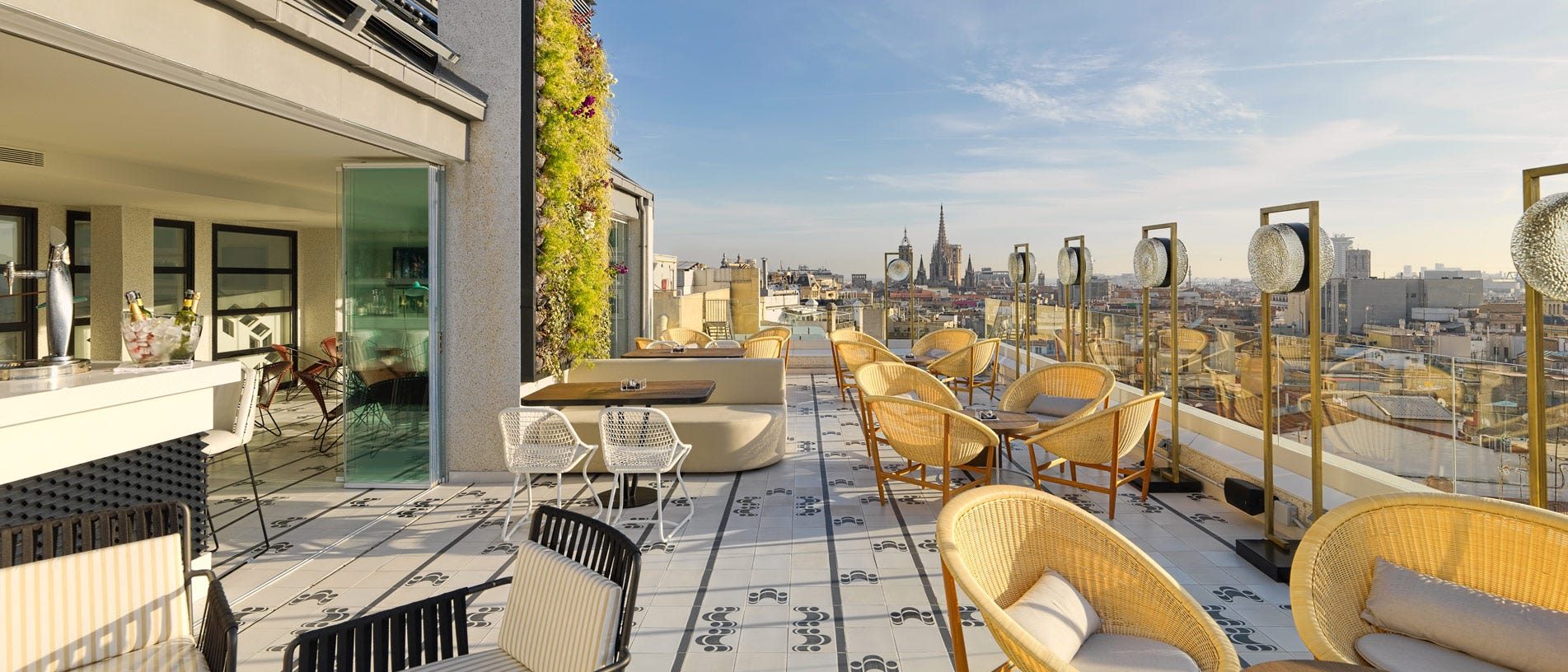 Atik rooftop terrace at H10 Cubik with wicker armchairs and low tables, overlooking Barcelona's rooftops toward the spires of the Sagrada Familia