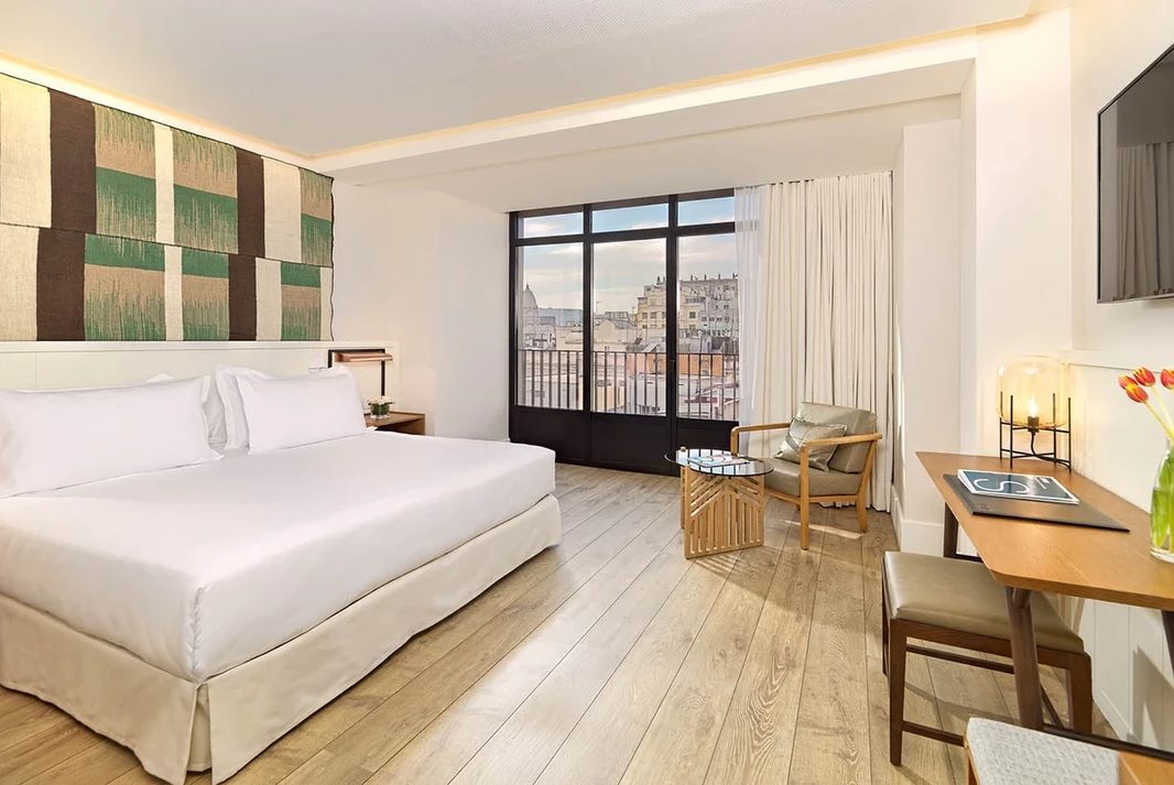 Superior room at H10 Cubik with a white bed against a green-and-brown patterned headboard panel and a Juliet balcony over the city