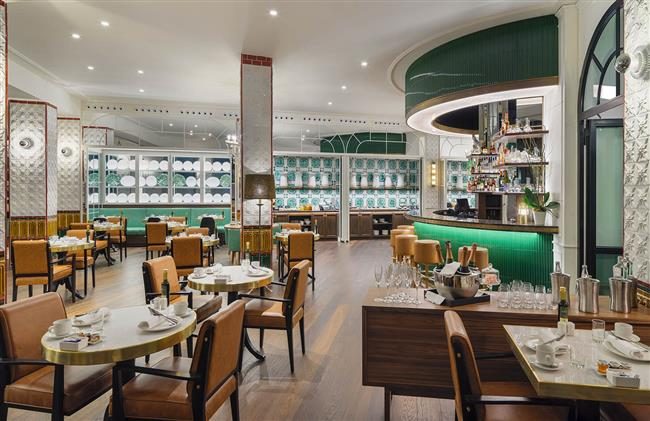 The Avenue Restaurant at H10 Madison with round marble tables, tan leather chairs and a curved green bar backed by shelves of blue-and-white patterned crockery