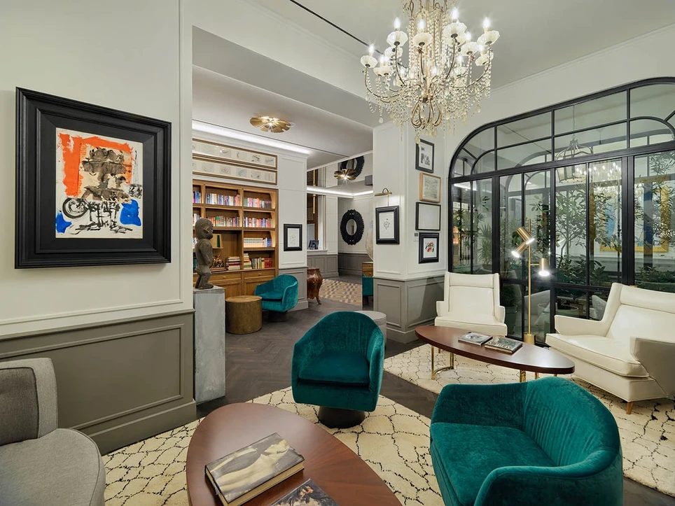 Lobby lounge at H10 Madison with teal velvet armchairs, a crystal chandelier and an abstract painting beside a glass-walled library nook