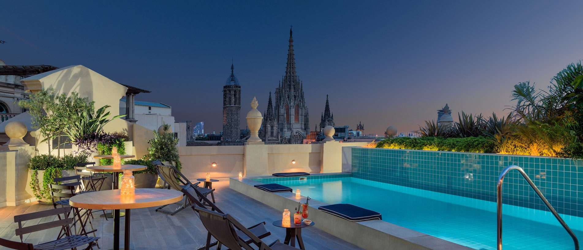 Rooftop pool at H10 Madison lit at dusk with cushioned loungers along the tiled edge and Barcelona Cathedral's spires rising behind