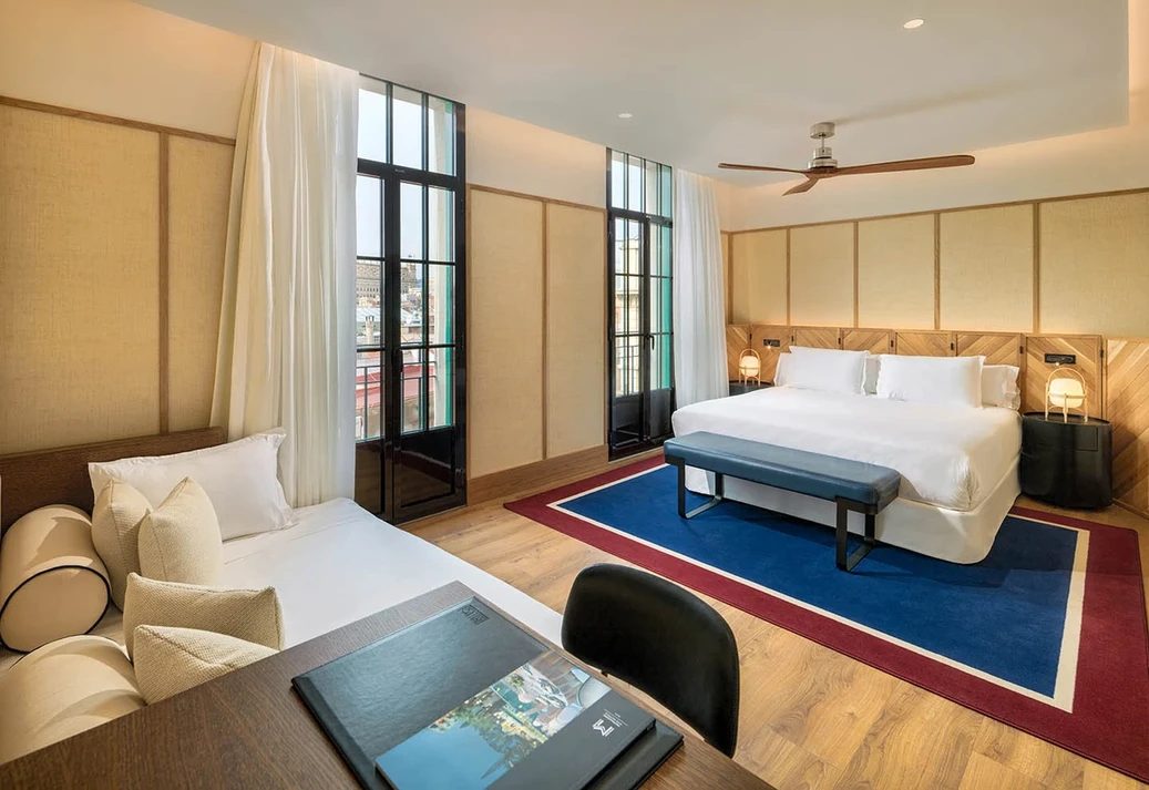 Junior Suite at H10 Madison with a white bed against a wood herringbone-panel headboard, a red-and-blue bordered rug and black-framed French doors onto a balcony