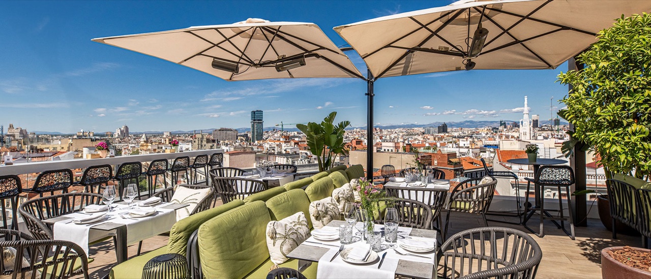 The El Cielo de Alcalá rooftop terrace set for dining under umbrellas, with green banquette seating and a panoramic view of Madrid's rooftops.