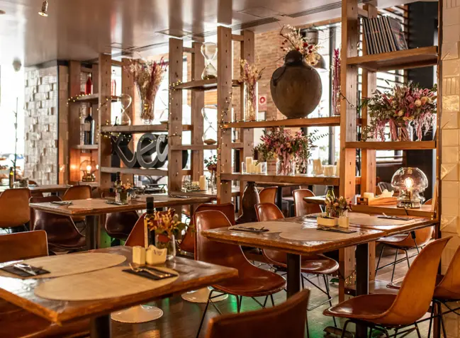 Tribeca Soho restaurant dining room with wooden tables, tan leather chairs and open shelving holding flowers and books.