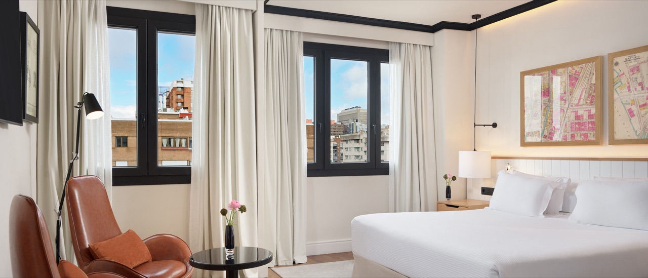Hotel bedroom with a white bed, tan leather armchair and floor-to-ceiling curtains framing city-view windows.