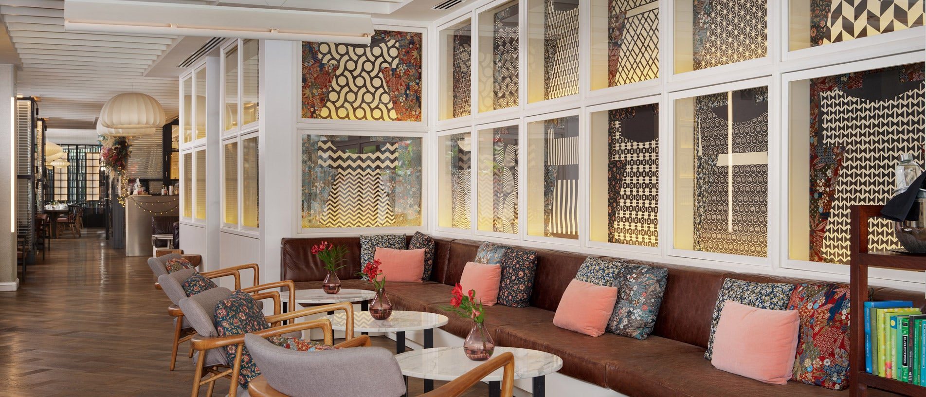 Lounge at H10 Universitat with a long brown leather banquette scattered with pink and patterned cushions beneath backlit geometric wall panels