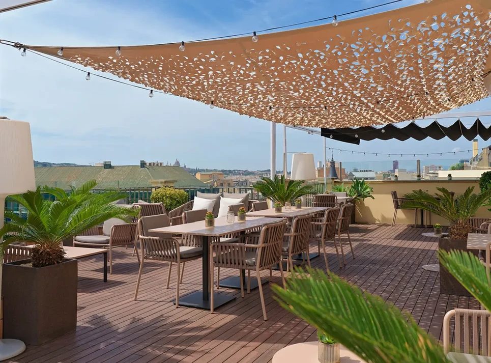 Twenty One rooftop terrace at H10 Universitat with wood decking, string lights, a perforated sail-shade canopy, and dining tables overlooking Barcelona