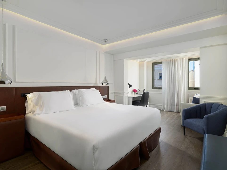 Junior suite at H10 Universitat with a white bed against a brown leather headboard and a separate seating alcove with a navy armchair by the window
