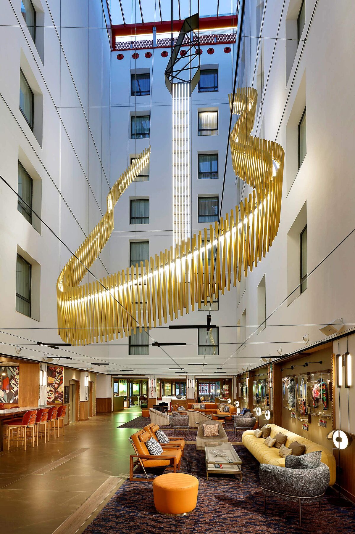 Tall hotel atrium lined with framed music memorabilia, a gold ribbon-shaped light sculpture suspended overhead between the guest-room floors.