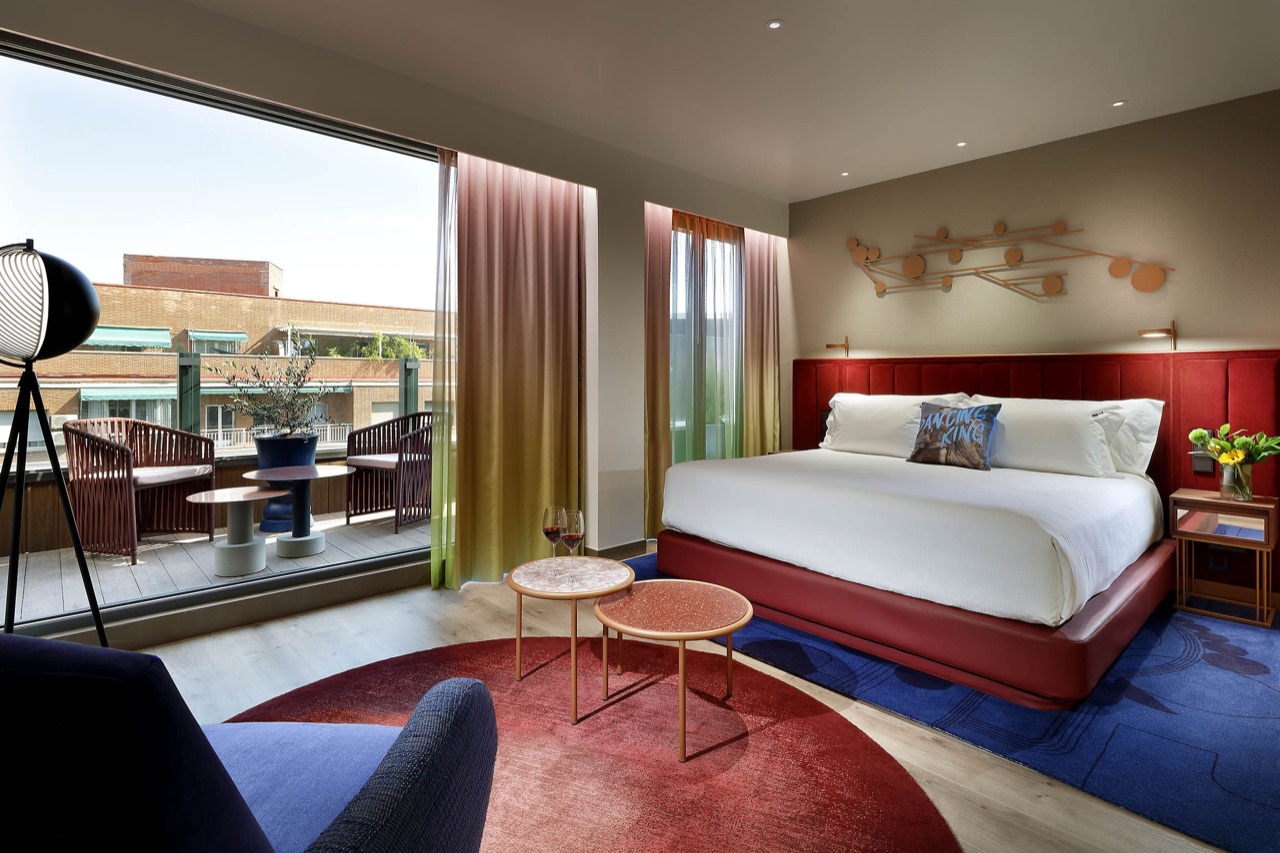 Hotel suite with a red upholstered bed facing a private terrace with rattan chairs, seen through a floor-to-ceiling glass door.