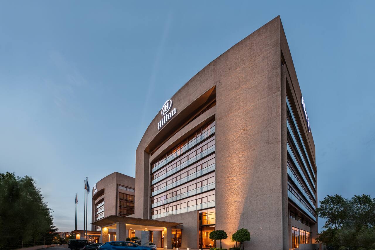 The angular concrete facade of Hilton Madrid Airport lit up at dusk, with the Hilton logo above the entrance canopy.