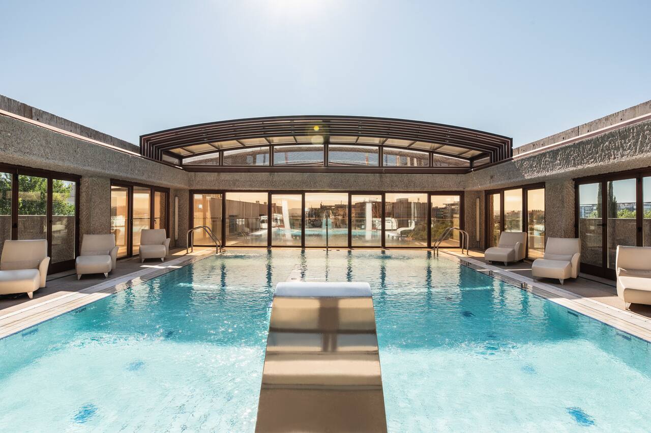 An indoor swimming pool with a retractable glass roof, a diving platform, and white lounge chairs along both sides.