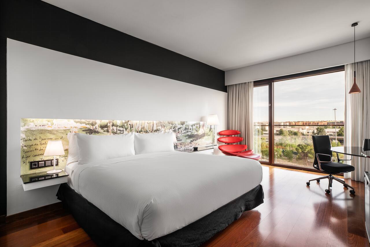 A king guest room with a white bed against a black accent wall with an illustrated headboard panel, a red lounge chair, and a floor-to-ceiling window.