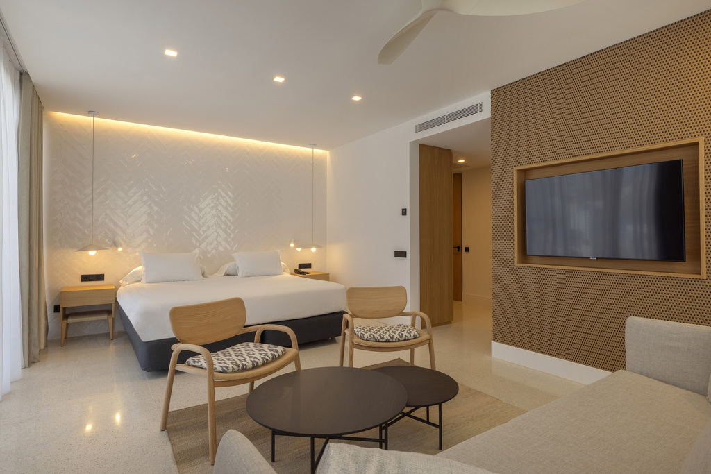 Double room at HM Palma Blanc in evening light, with a wall-mounted TV in a perforated wood panel and two chairs by the bed.