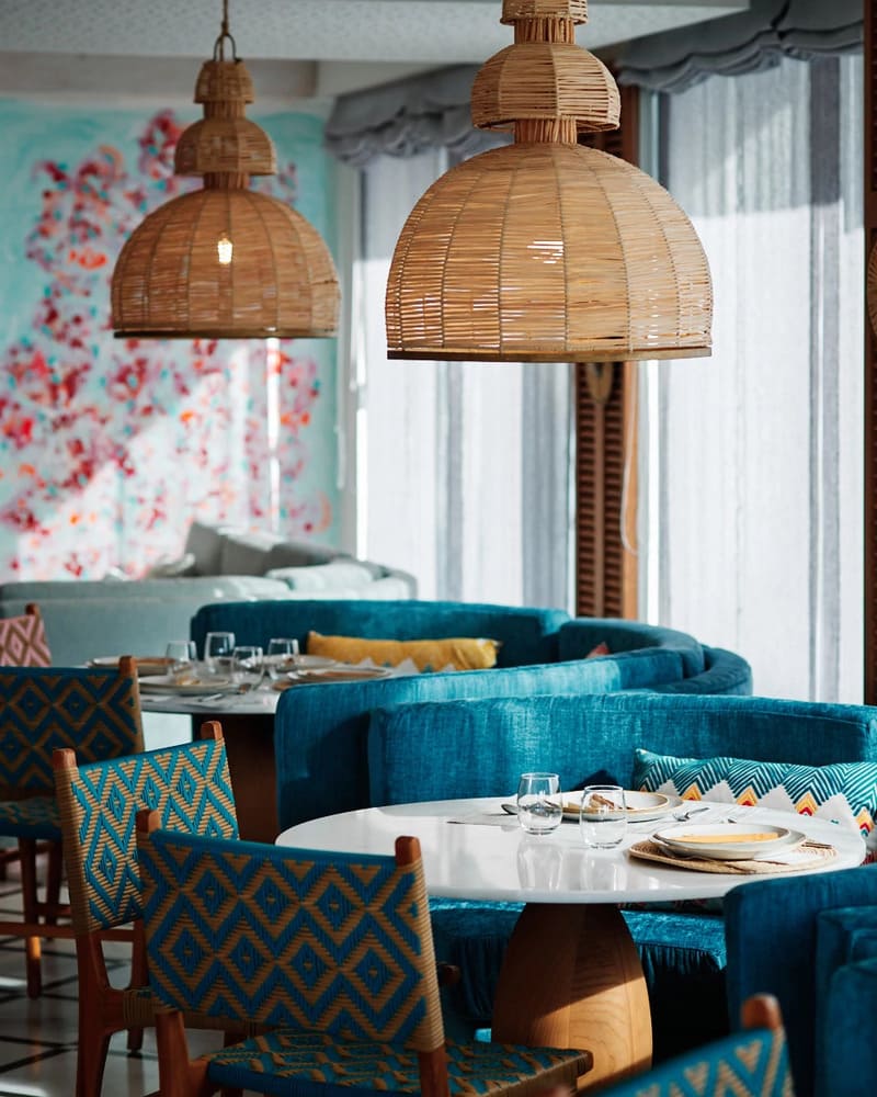 The dining room of Restaurant Salicornia at Hotel Honucai, with rattan pendant lamps, blue banquette seating and round white tables.