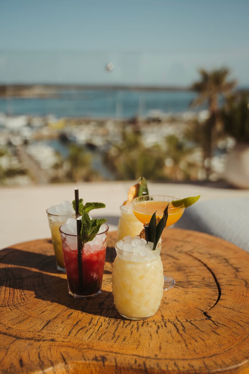 Four cocktails on a wooden table at The Top rooftop bar, with the marina and palm trees blurred in the background.