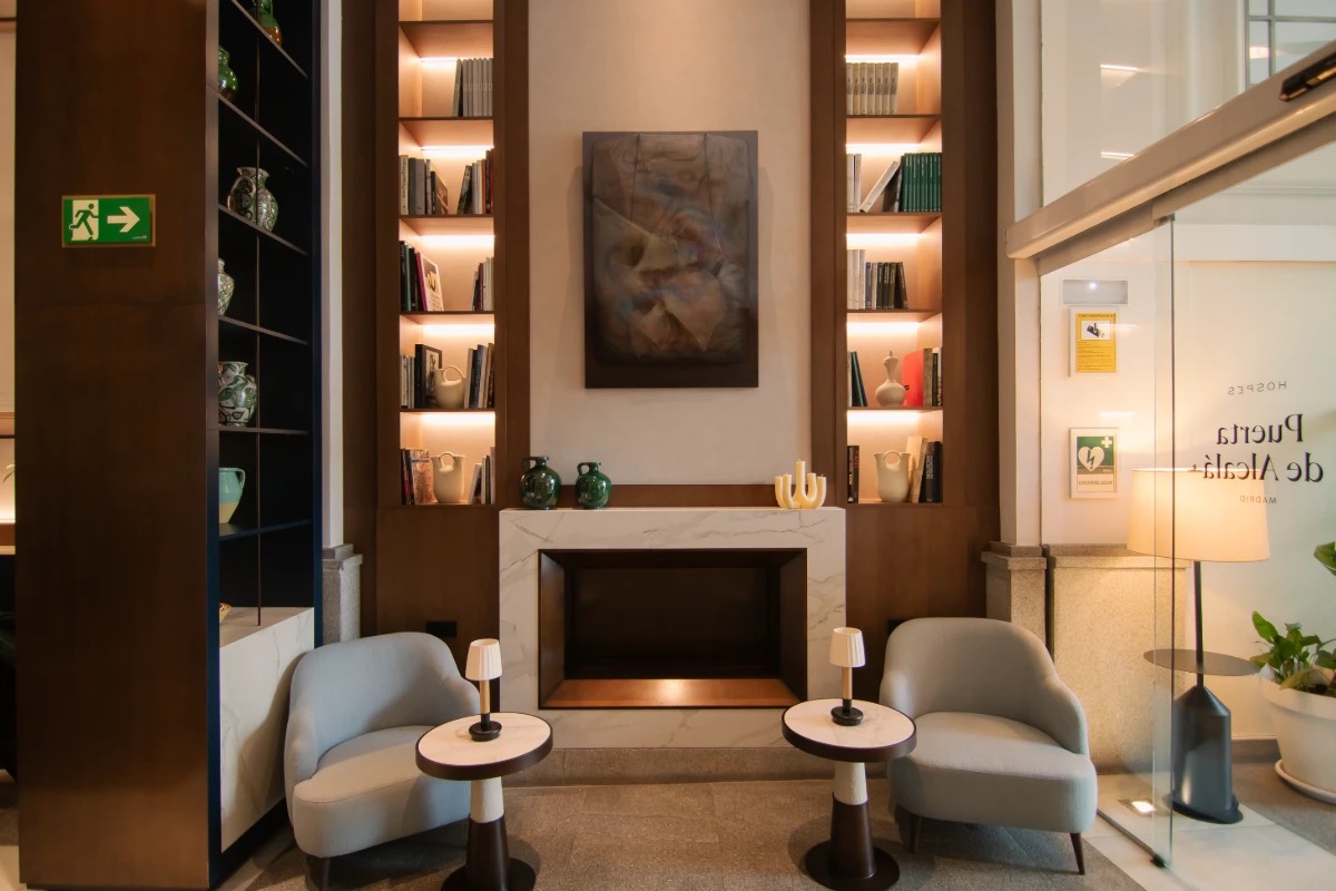 A lobby lounge with a white marble fireplace between backlit wooden bookshelves holding books and ceramic jugs, two pale blue armchairs with small round tables, and a glass entrance door lettered Hospes Puerta de Alcala on the right.