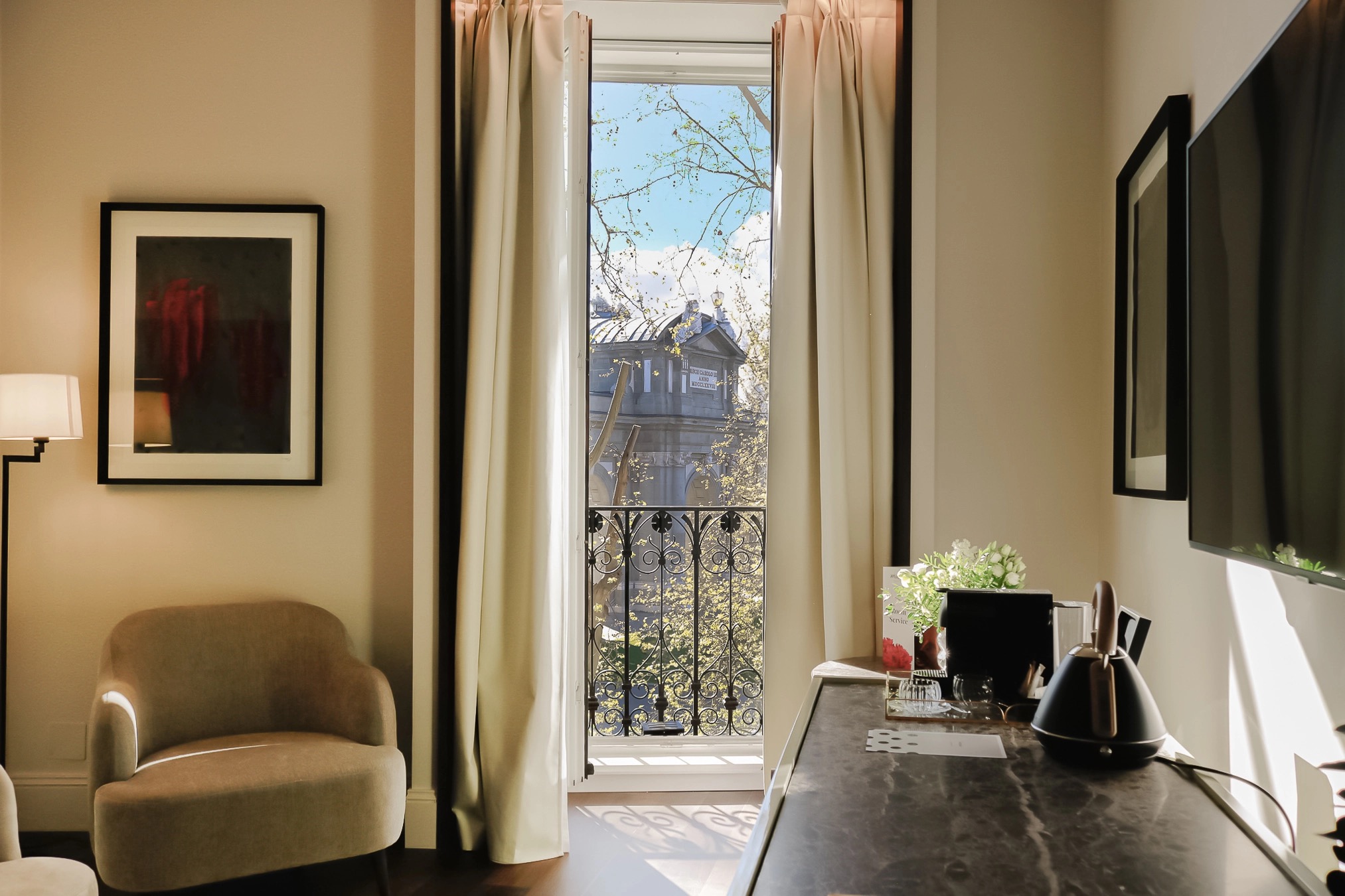 A guest room with a beige armchair, a framed red abstract print, and an open French window with a wrought-iron balcony looking straight onto the Puerta de Alcala gate; a dark marble desk with a kettle and coffee machine stands on the right.