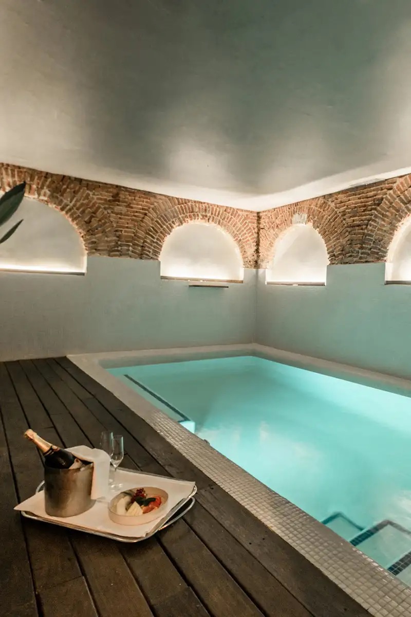 A small indoor spa pool with pale turquoise water under a whitewashed vault with exposed brick arches, a dark timber deck in the foreground and a tray with an ice bucket and glasses at its edge.