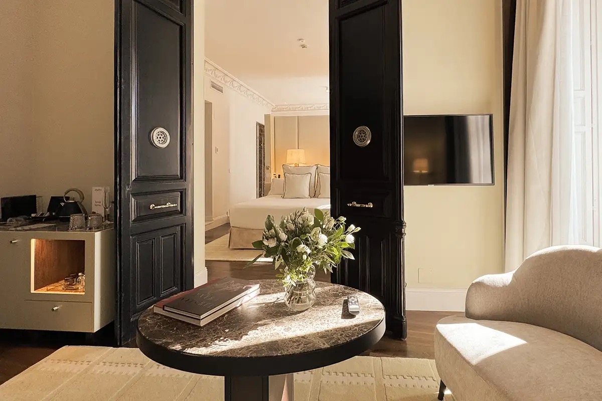 A suite sitting area with a round dark marble table holding books and a vase of white flowers, a cream curved sofa, a minibar cabinet, and tall black panelled double doors opening onto a bedroom with a bed and table lamps.