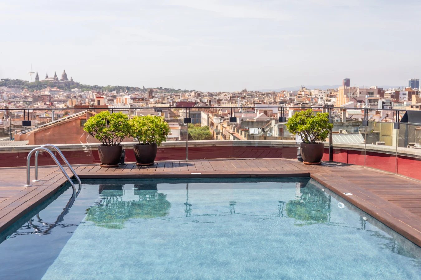 Rooftop pool at Hotel 1898 with wood decking and potted trees, overlooking Barcelona's rooftops toward Montjuic