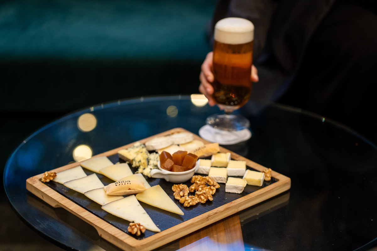 A wooden cheese board with sliced cheeses, walnuts, and quince paste on a napkin printed with the abba logo, next to a glass of beer.