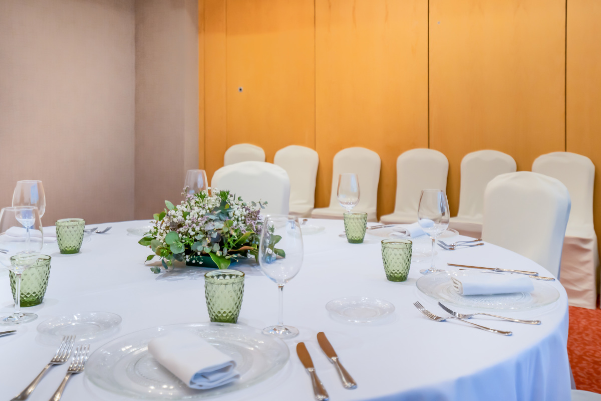A round banquet table set with white linens, glassware, folded napkins, and a floral centrepiece, surrounded by chairs with cream slipcovers.