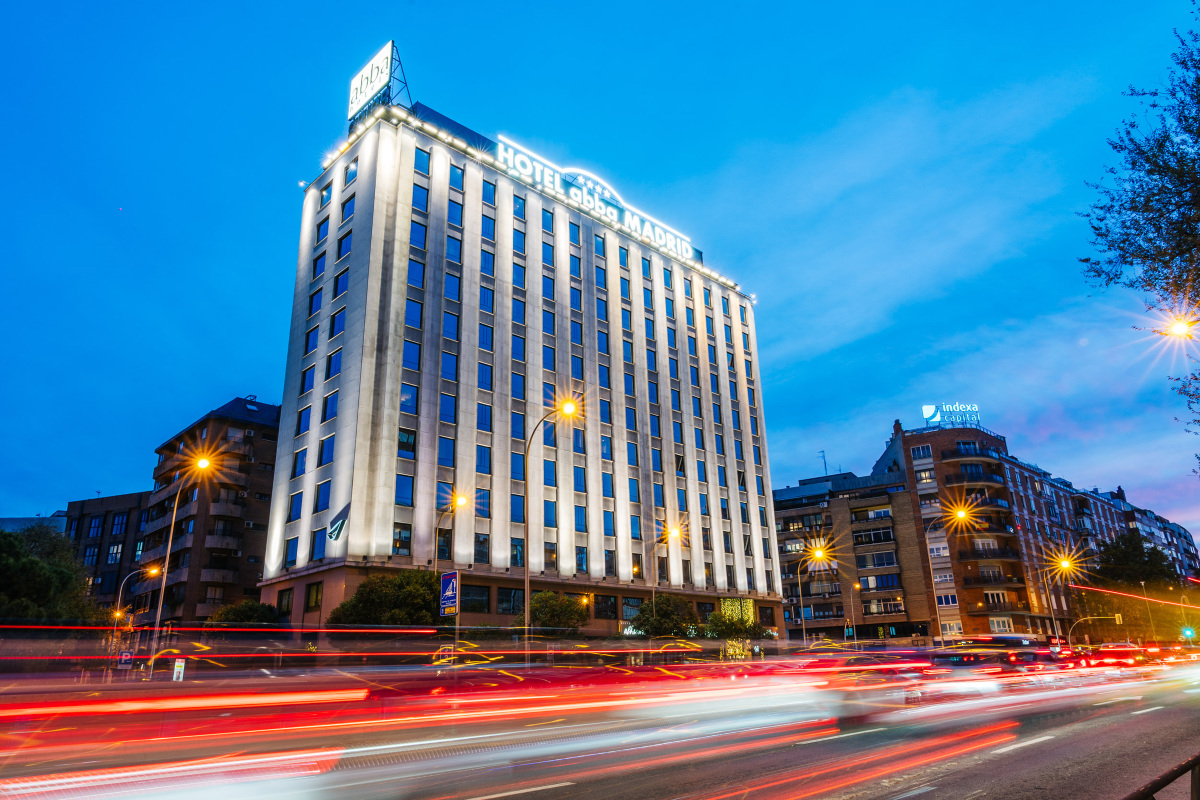 The hotel's lit tower facade at night with 'HOTEL abba MADRID' signage above illuminated vertical fins, over a busy avenue with light-trail traffic.