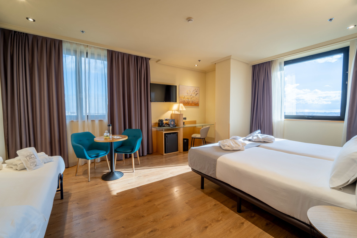 A Family room with two beds, a small seating area with teal chairs and a round table, and a wall-mounted TV.