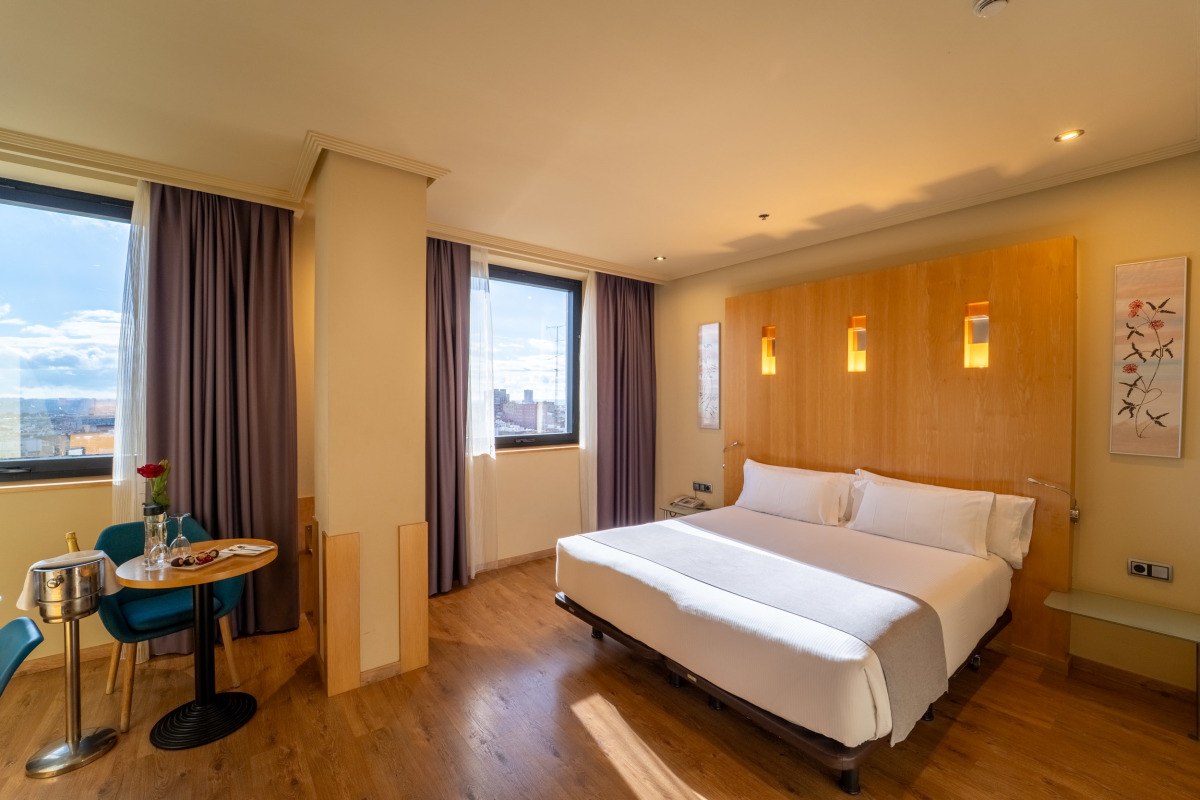 A Deluxe room with a wood headboard wall, white bedding, a small round table with two teal chairs, and a window looking over the city.