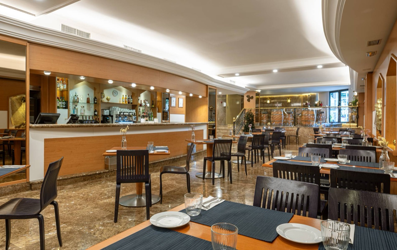 The bar and cafe area at Hotel Agumar, with a wood bar counter, glass shelving and small dining tables set with glasses.