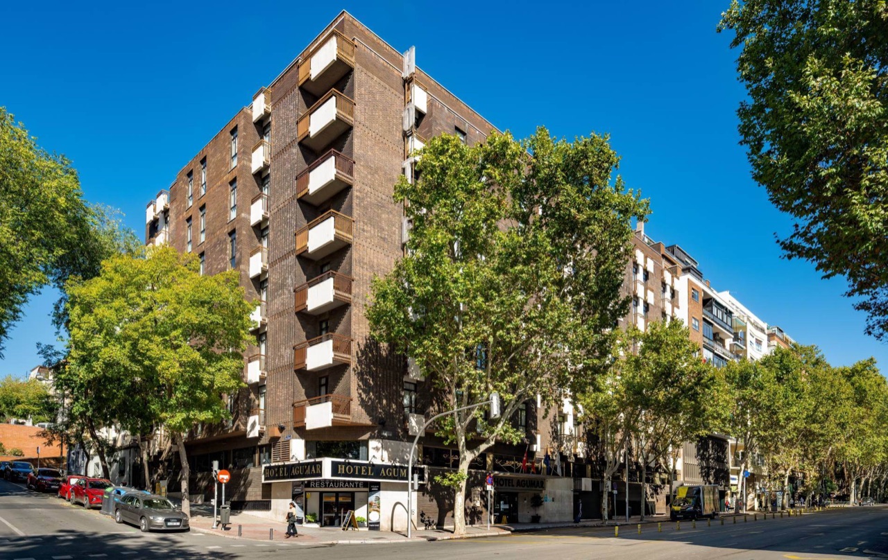 The brick facade of Hotel Agumar on a tree-lined Madrid avenue, with the hotel's name in gold letters above the entrance.