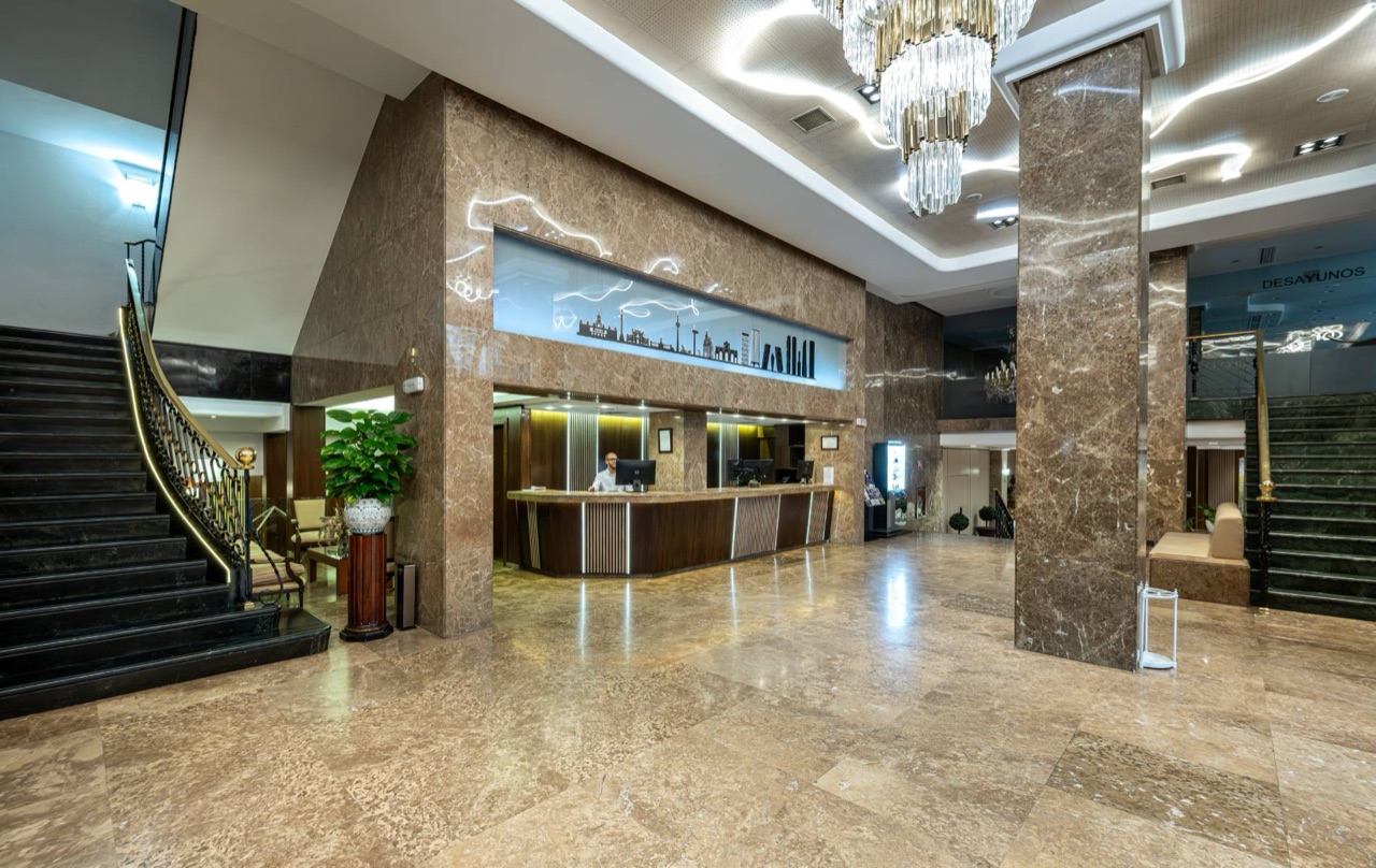 The marble-floored lobby of Hotel Agumar, with a curved reception desk, a crystal chandelier and a black staircase with brass railing.