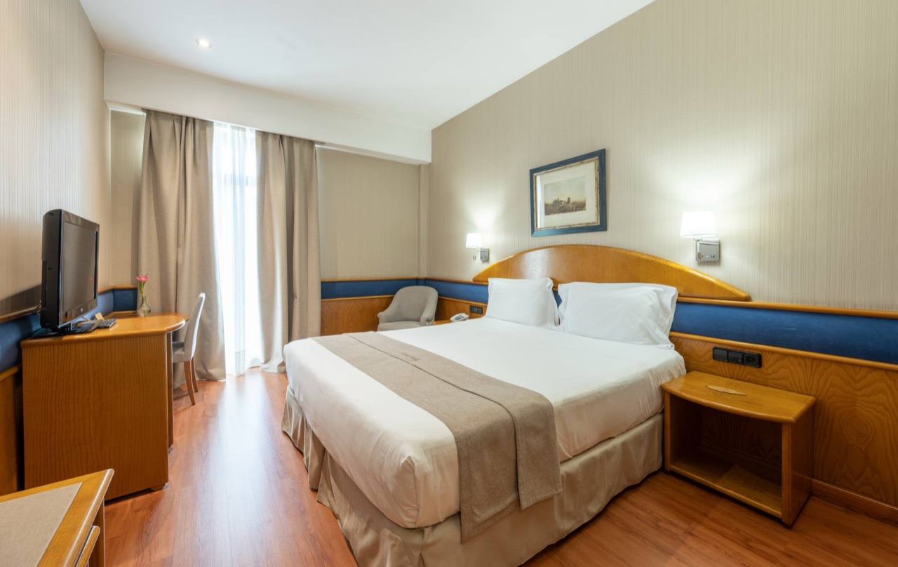 A hotel double room with a wood headboard, white and taupe bedding, a desk with a TV, and curtained balcony doors.