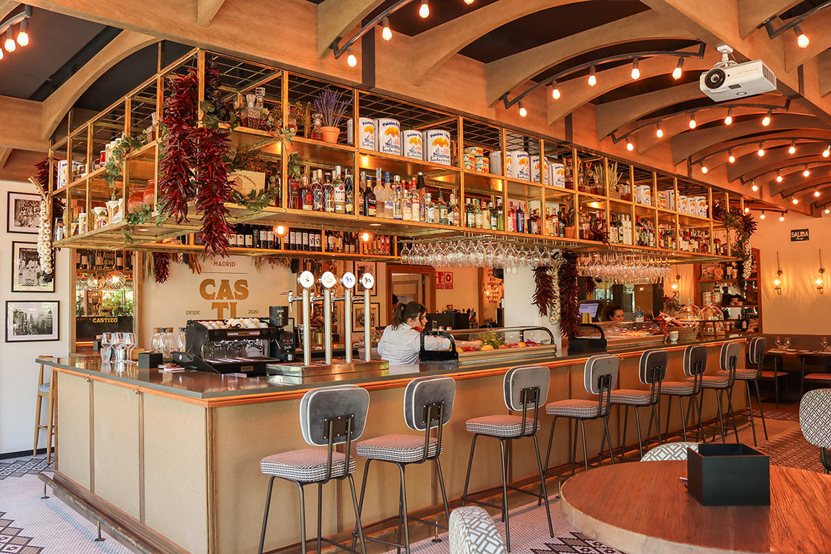 The Castizo bar counter with a shelf of bottles, hanging wine glasses and dried red peppers, and a 'Madrid Castizo desde 2020' sign on the back wall.