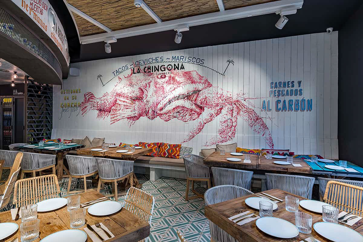 The La Chingona dining room, with wooden tables set for two and a large red mural of a crustacean above a banquette reading 'La Chingona'.