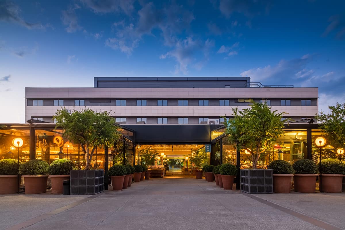 The hotel building at dusk, seen from a courtyard lined with potted trees, with the glass-fronted Rumbo Aravaca restaurants lit up at ground level.