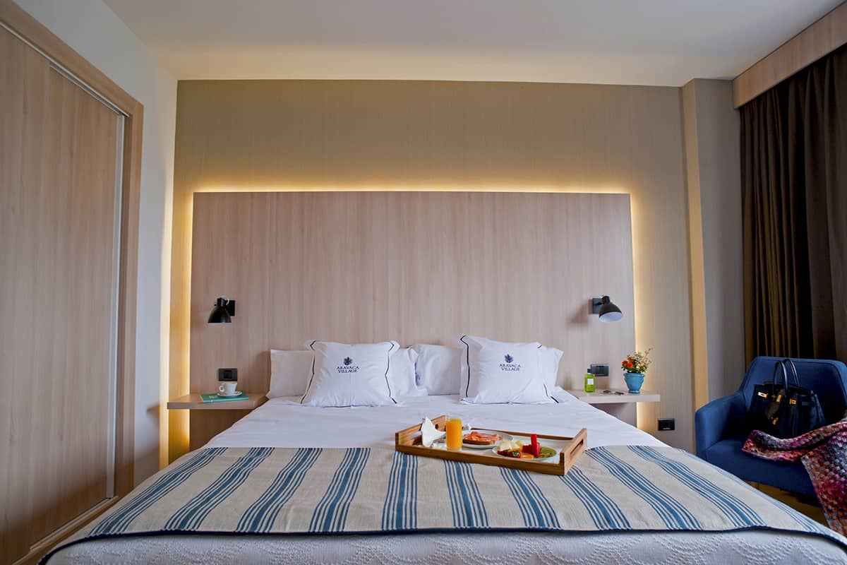 A hotel bed with Aravaca Village branded pillows and a wooden breakfast tray holding orange juice and pastries.