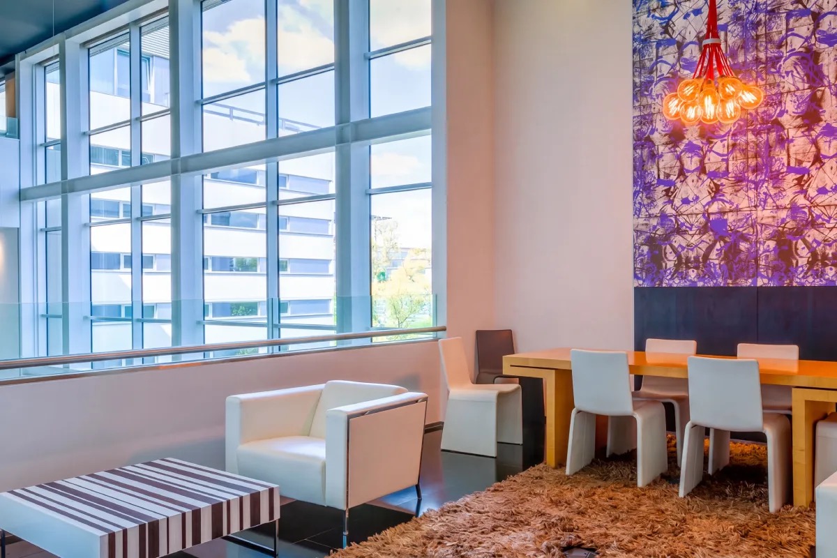 Bright dining or lounge area with a wooden table and white chairs on a shaggy rug, floor-to-ceiling windows and a purple patterned wall panel with a pendant light.