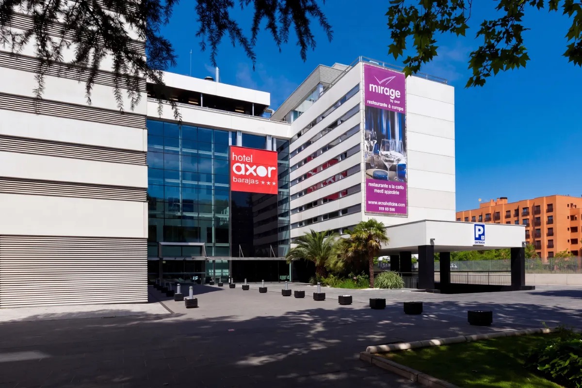 White multi-storey hotel building with a red Hotel Axor Barajas sign and a purple Mirage by Axor restaurant banner on the facade, palm trees in front.