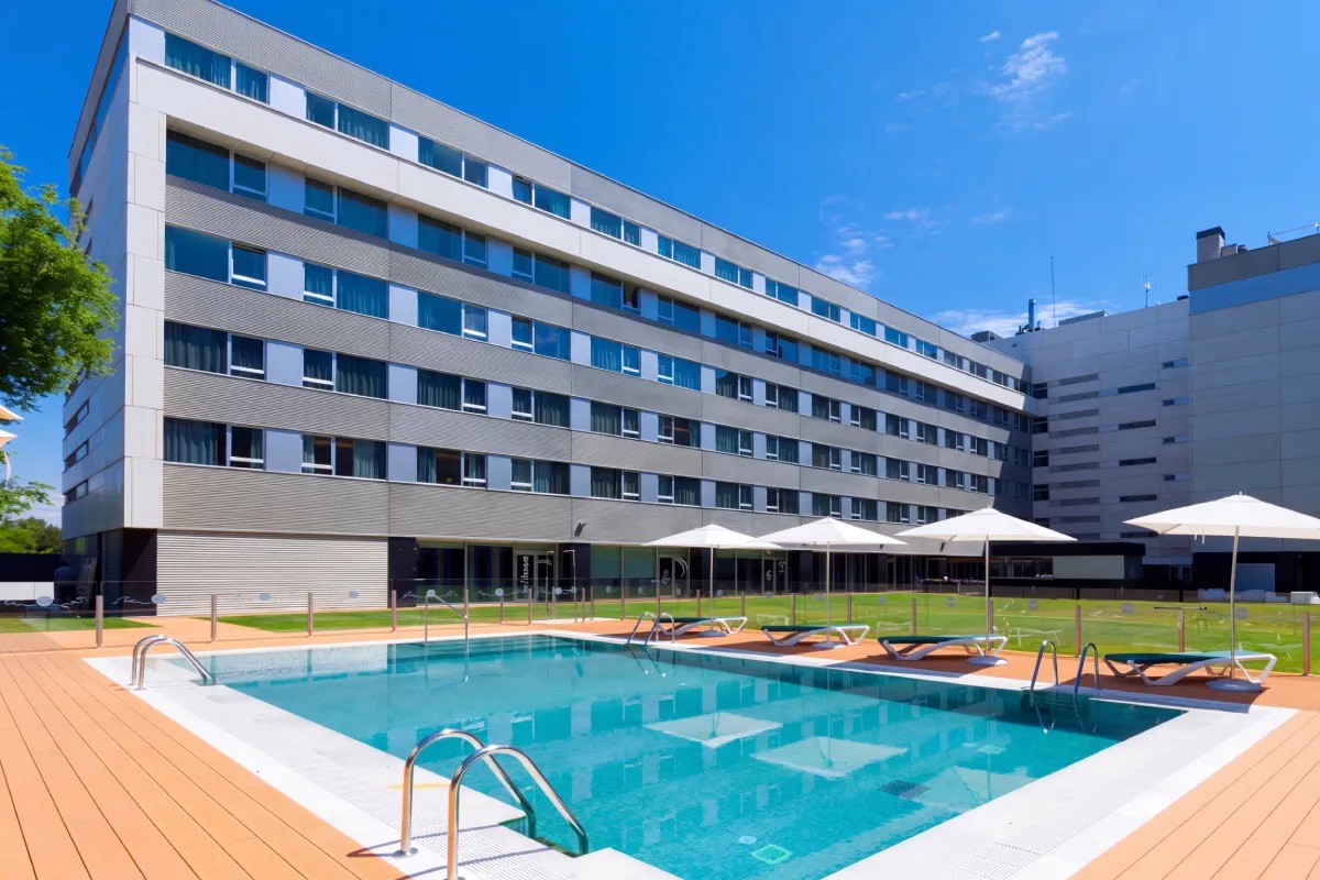Outdoor swimming pool with a wooden deck and white sun loungers under umbrellas, set against the hotel's grey facade and a lawn.